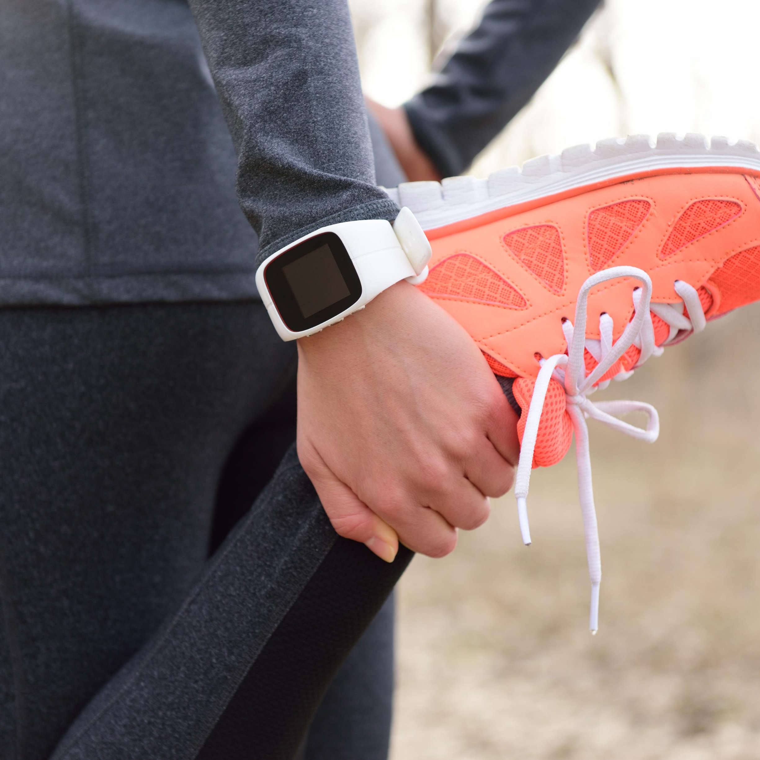 Individual Stretching front of leg with grey long sleeve top, black leggings, white watch and coral sneakers, represents “Feeling Sluggish, 30 Min Class to Jumpstart Your System” program