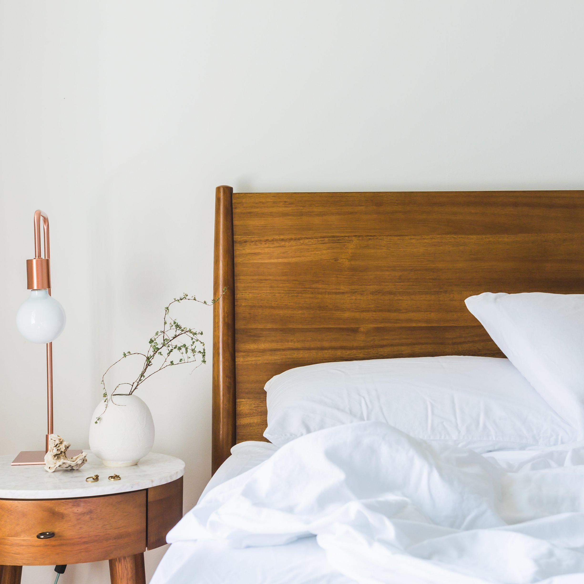 Bed with wood headboard, white sheets, wooden night stand with white vase and lamp against a white wall, represents “Need Some Sleep Advice?” program